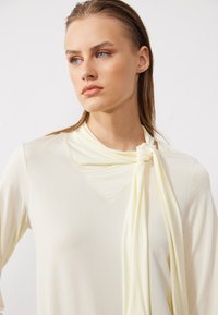 Cremfarbener langärmliger Oberteil mit V-Ausschnitt und drappiertem Schalsdetail, das am Hals gebunden ist, aus glattem Stoff mit weicher Textur.