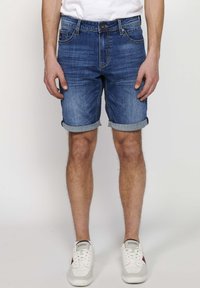 Pantalones cortos de mezclilla en un color azul medio con un dobladillo enrollado, que presentan bolsillos en la parte delantera y trasera, y un sutil patrón de decoloración. Llevados con zapatillas blancas.