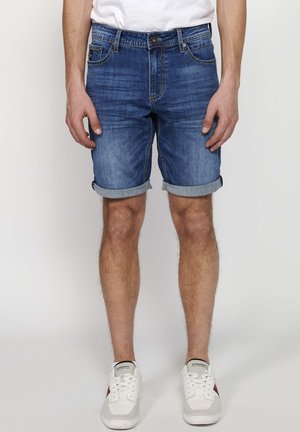 Keskmise sinise värvi denim lühikesed püksid, rullitud äärtega, ees ja taga taskud, kerge pleekimisega muster. Kandnud valgeid spordijalatsid.