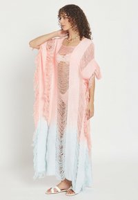 Mujer con cabello rizado que lleva una cubierta larga de flecos rosa y blanca sobre un vestido desgarrado, de pie con una mano cerca del cuello.