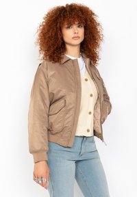Lockenhaarige Frau, die eine hellbraune Bomberjacke, eine cremefarbene Knopfleiste, hellblaue Jeans und silberne Ringe an der Hand trägt, vor einem weißen Hintergrund.