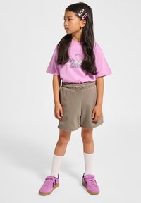 Rosa t-shirt med grafisk text, beige shorts och rosa sneakers med mörka accenter. Långt mörkt hår med klämmor, stående pose.
