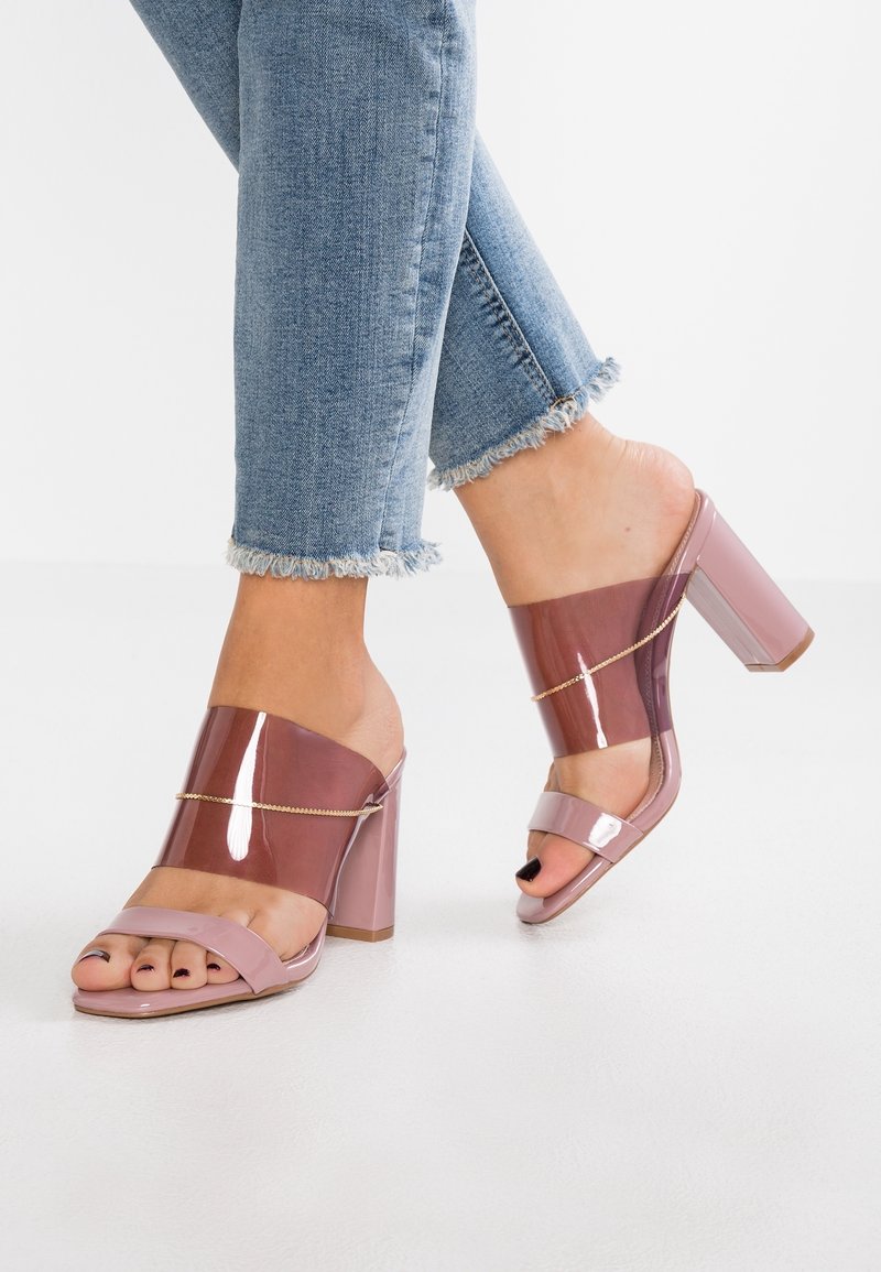 Mules à talons bloc roses avec des straps transparents, dotées d'un accent en chaîne dorée et d'une finition lisse et brillante. Jean avec des ourlets effilochés.