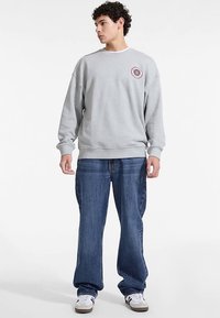 Grijze trui met een ronde hals en een ronde embleemdetail. Draagt losse blauwe jeans en witte sneakers, wat een casual uitstraling geeft.