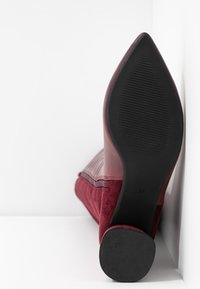 Botte à talon haut bordeaux avec un bout pointu, présentant une tige lisse avec des coutures verticales, un côté en suède texturé et une semelle noire.