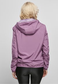 Blondi nainen kääntyy pois violetissa hupparikaupassa ja mustissa ribattuissa leggingsseissä tasaisella harmaalla taustalla.