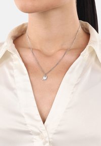 Närbild av en kvinna som bär en oformad krämfärgad blus och ett silversmycke med ett "M"-hänge runt halsen.