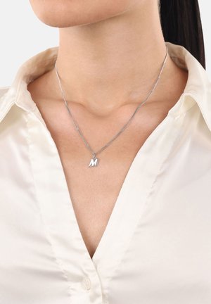 Nahaufnahme einer Frau, die eine ungeknöpfte cremfarbene Bluse und eine silberne Halskette mit einem "M"-Anhänger um den Hals trägt.