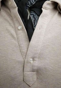 Lys beige skjorte med krave i tekstureret stof, der har en tre-knap lukning. Et sort mønstret tørklæde kan ses under kraven.