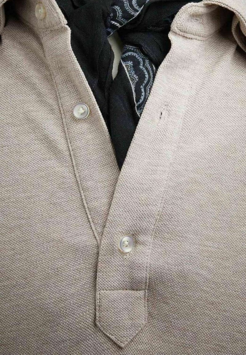 Lys beige skjorte med krave i tekstureret stof, der har en tre-knap lukning. Et sort mønstret tørklæde kan ses under kraven.