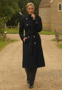Dug crni vuneni kaput sa zlatnim dugmadi, visokim ovratnikom i pojasom, u kombinaciji s crnim hlačama i čizmama na petu; postavljen protiv prirodne vanjske pozadine.