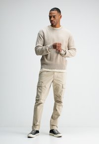 Beiger gerippter Pullover kombiniert mit hellbeigen Cargo-Hosen. Bemerkenswerte Details sind seitliche Taschen und schwarze Sneaker mit weißen Sohlen.