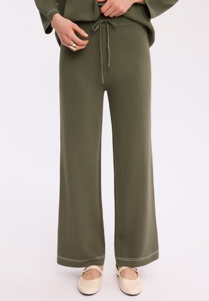 Osoba nosząca oliwkowo zielone szerokie spodnie ze ściągaczem w pasie i beżowe płaskie buty z paskiem i klamrą.