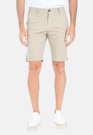 Uomo che indossa pantaloncini beige fino al ginocchio, scarpe da ginnastica bianche e camicia bianca, in piedi con le mani rilassate ai lati.