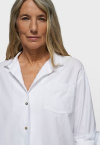 White button-up shirt made of textured fabric, featuring a collar, a single chest pocket, and rolled-up sleeves.