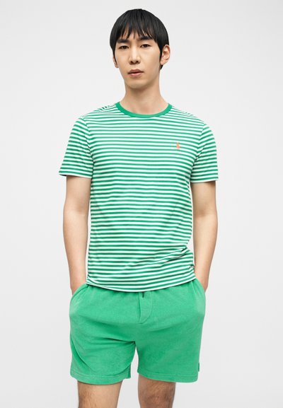 Άνδρας που στέκεται με τα χέρια στις τσέπες φορώντας πράσινη και λευκή ριγέ μπλούζα και μονόχρωμο πράσινο σορτς, σε απλό ανοιχτό φόντο.