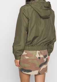 Giacca bomber leggera color verde oliva con cappuccio, polsini e vita elasticizzati, abbinata a una gonna con motivo camouflage in toni terrosi.