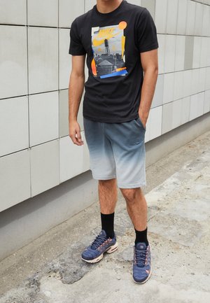Mężczyzna stojący na zewnątrz, ubrany w czarną koszulkę z grafiką Nike, niebiesko-szare szorty ombre, czarne skarpetki i niebieskie buty sportowe.