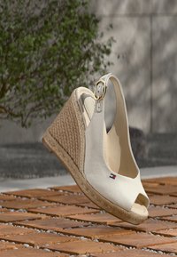 Λευκό υφασμάτινο πέδιλο με πλατφόρμα σε σφήνα, ανοιχτή μύτη και σχεδιασμό με λουράκι στο πίσω μέρος. Διαθέτει πλεκτή φτέρνα από σκοινί λινάτσας και χρυσή αγκράφα ως διακοσμητικό στοιχείο.