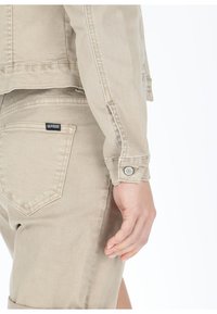 Beige Denimjacke mit geknöpften Bündchen, von der Seite gezeigt, das die Tasche hervorhebt. Das Etikett "SEASIDE" ist auf der Rückentasche sichtbar.