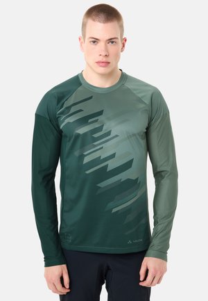 Jeune homme portant un maillot de sport à manches longues vert foncé avec des motifs géométriques et un pantalon noir, faisant face à l'avant sur un fond blanc.