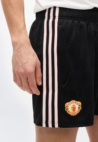 Δεξί χέρι δίπλα σε μαύρο αθλητικό σορτς με κάθετες λευκές και κόκκινες ρίγες και ένα πιετάκι με το λογότυπο της Μάντσεστερ Γιουνάιτεντ κοντά στο τελείωμα.