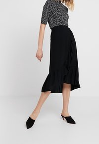 Falda negra con dobladillo con volantes, combinada con una blusa de lunares en blanco y negro. Modelo usando mules negros se encuentra sobre un fondo neutro.