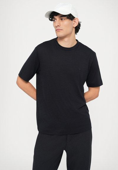 Czarny t-shirt z krótkim rękawem i okrągłym wycięciem przy szyi, ozdobiony subtelnym logo. Połączony z ciemnymi spodniami, na jednolitym białym tle.