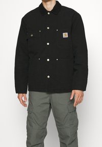 Czarna kurtka z płótna z zatrzaskami, dwoma kieszeniami na piersiach i kołnierzem z sztruksu. Nosi się na szare spodnie cargo dla praktycznego wyglądu.
