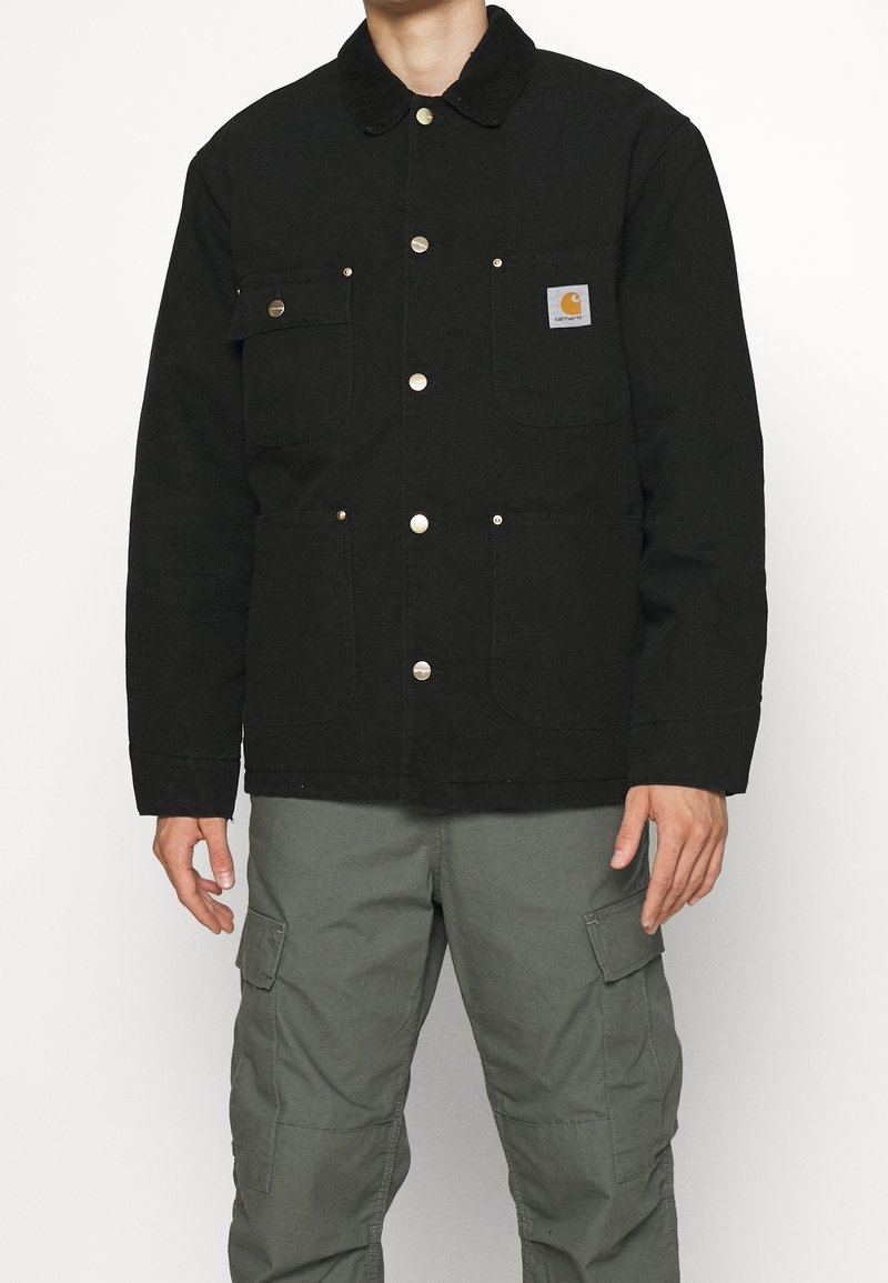 Czarna kurtka z płótna z zatrzaskami, dwoma kieszeniami na piersiach i kołnierzem z sztruksu. Nosi się na szare spodnie cargo dla praktycznego wyglądu.