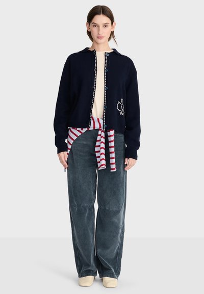 Jeune femme portant un cardigan bleu marine avec une feuille brodée, un pull rayé noué à la taille, un pantalon large gris et des chaussures crème, debout sur fond blanc.