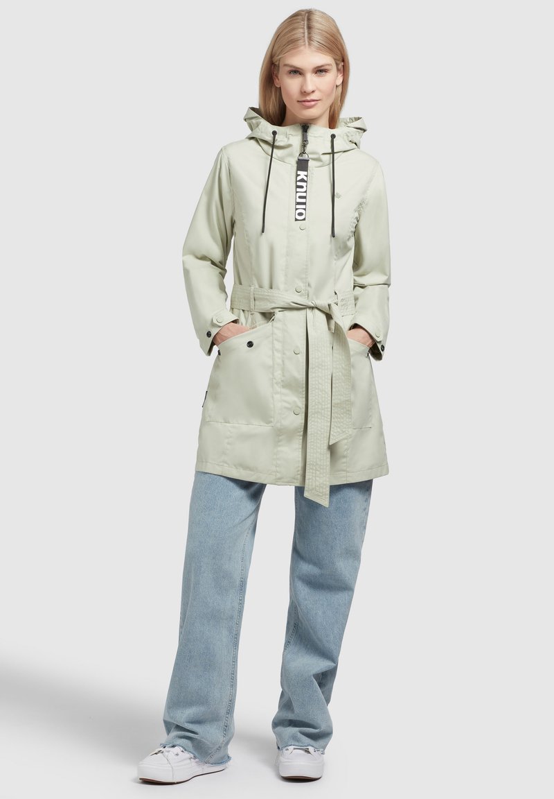 Giacca impermeabile verde chiaro con cappuccio regolabile, vita con cintura, bottoni a pressione e due tasche frontali, abbinata a pantaloni in denim chiaro.