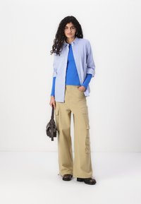 Chemise cintrée à rayures bleu clair sur un t-shirt bleu, associée à un pantalon cargo beige à jambes larges. Les accessoires comprennent un petit sac à main sombre.