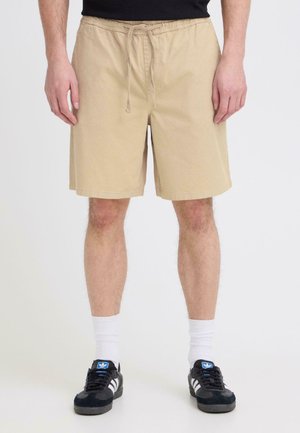 Mand iført beige knælange shorts med snøre, hvide sokker og sorte Adidas-sneakers, stående mod en ensfarvet baggrund.