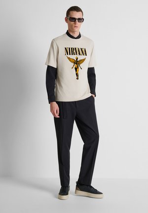 Joven con gafas de sol negras, camiseta beige de Nirvana sobre mangas largas negras, pantalones negros y zapatillas negras con suelas beige, de pie.