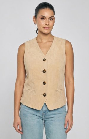 Kvinde iført ærmeløs beige knappet vest og lyseblå jeans står mod en ensfarvet lys baggrund.