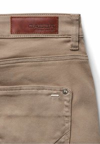 Bézs színű vászonnadrág barna bőrcímkével a deréknál. Cipzáras zsebbel és fém kiegészítőkkel rendelkezik. Tartós kivitel.