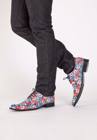Bloemdruk schoenen in blauw, rood en wit met een glanzende afwerking. Lage hak, spitse neus en zwarte veters. G gedragen met donkere jeans.