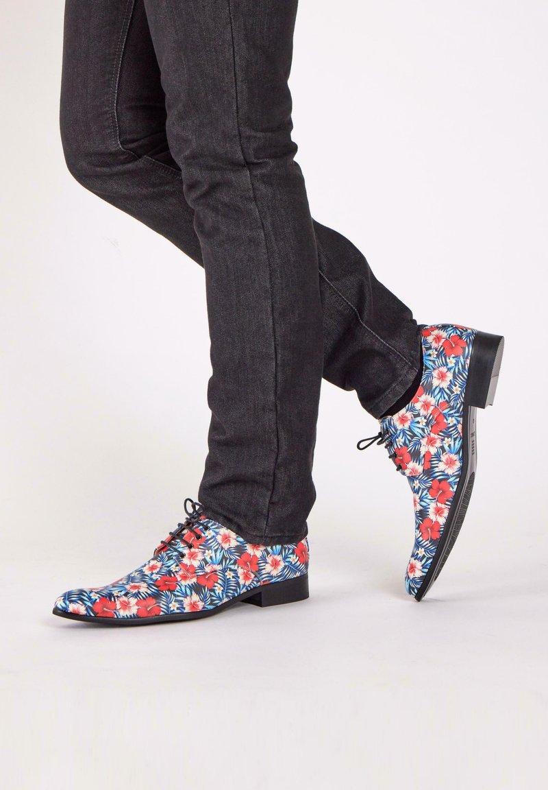 Bloemdruk schoenen in blauw, rood en wit met een glanzende afwerking. Lage hak, spitse neus en zwarte veters. G gedragen met donkere jeans.
