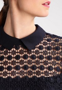 Navy-Bluse aus Spitze mit einem strukturierten Stoff, floralen Mustern und einem klassischen Kragen. Texturierte Details erhöhen das visuelle Interesse.