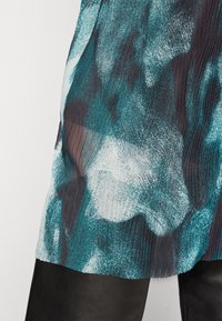 Haut transparent plissé affichant un motif abstrait dans des teintes de bleu-vert, noir et blanc ; texture lisse avec un design fluide.