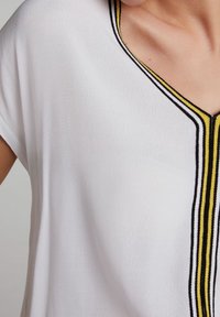 Top blanco con escote en V, que presenta una tela texturada y acentos a rayas en negro y amarillo a lo largo del escote. Mangas cortas y holgadas.
