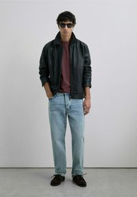 Chaqueta de cuero negra con cremallera, camiseta granate, vaqueros rectos de un azul claro y zapatos de ante marrón oscuro. Unas gafas de sol redondas completan el look.