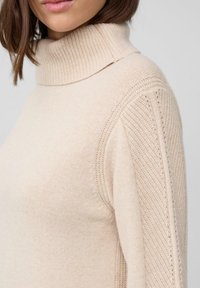 Beżowy sweter z golfem, wykonany z miękkiego materiału dzianinowego, z prążkowaną teksturą przy kołnierzu i dekoracyjnymi wzorami dzianinowymi wzdłuż rękawów.