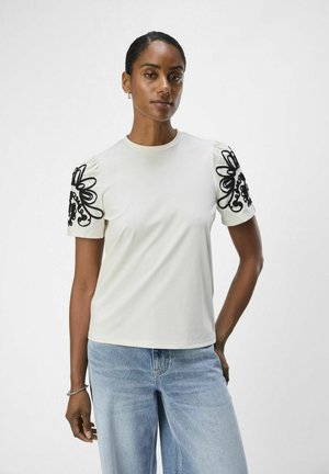 Kortärmad krämfärgad T-shirt med svart blommönstrad broderi på puffärmarna. Slät tyg, avslappnad passform, kombinerad med ljusblå jeans.