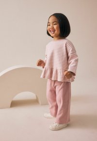 Pantaloni rib generați roz deschis și un top cu un model roz și alb, cu un tiv de tip peplum. Copilul poartă adidași albi și zâmbește.