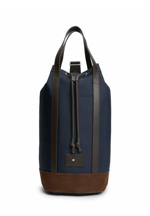 Granatowy plecak ze ściągaczem, brązową zamszową podstawą, ciemnymi skórzanymi paskami i małą prostokątną naszywką z logo z przodu.