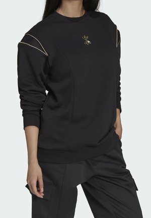 Vrouw draagt een zwart sweatshirt met gouden bies op de schouders en een goudkleurig logo op de borst, gecombineerd met zwarte cargobroeken.