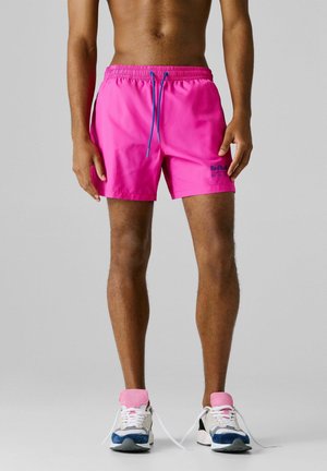 Mann trägt leuchtend pinke Red Bull Racing Shorts und weiße Sneakers mit blauen und pinken Akzenten, steht vor einem grauen Hintergrund.