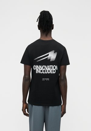 Mann mit Dreadlocks, der ein schwarzes T-Shirt mit weißem Text "INNOVATION INCLUDED" und einem Grafikdruck auf der Rückseite trägt, kombiniert mit lockeren grauen Hosen.